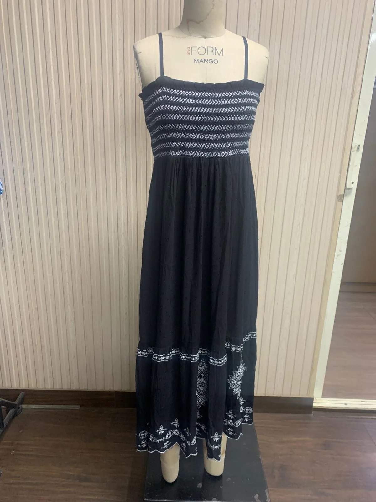 Front view of a black smocked strappy maxi dress with embroidered hem on a dress form